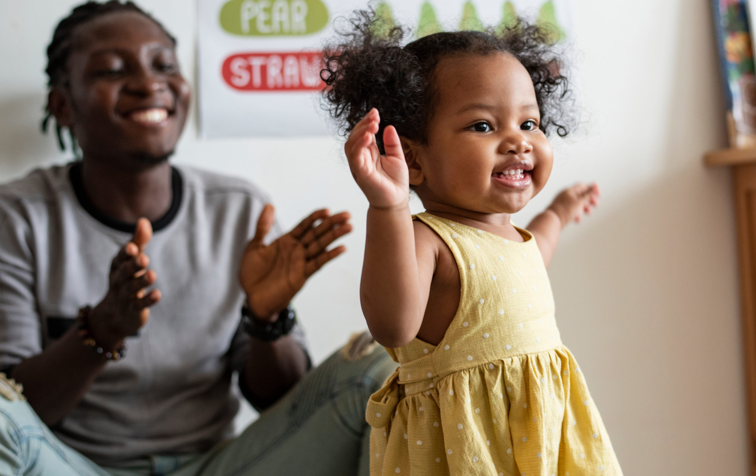 A child in a yellow dress joyfully raising their hands, with a man clapping in the background